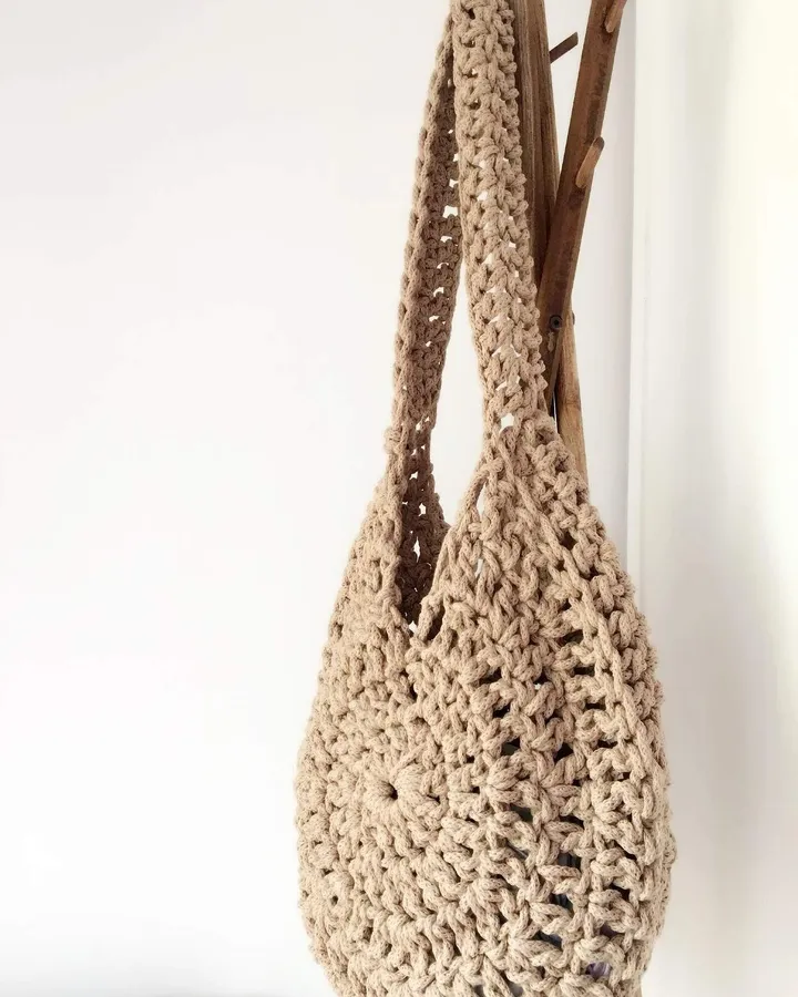 Bolso de crochet en camel, colgado de un perchero de madera: el punto cede y el cuerpo se alarga