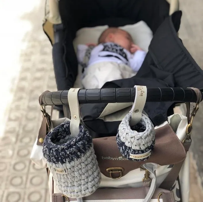 Dos portabibis colgados del manillar de un cochecito, uno con el nombre Noah, en un paseo por la ciudad