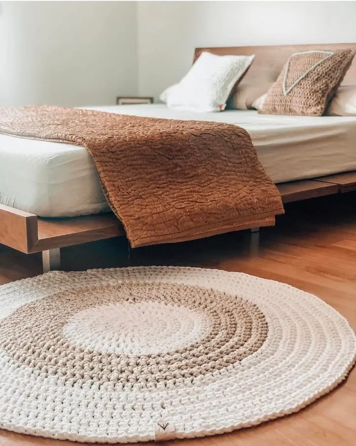 Alfombra redonda de crochet en crudo y arena a los pies de la cama, con el cojín del triángulo sobre las almohadas
